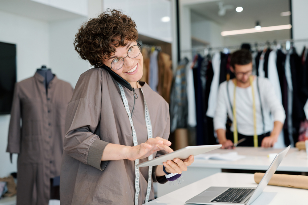 A woman on the phone while working on a laptop and tablet in a tailoring